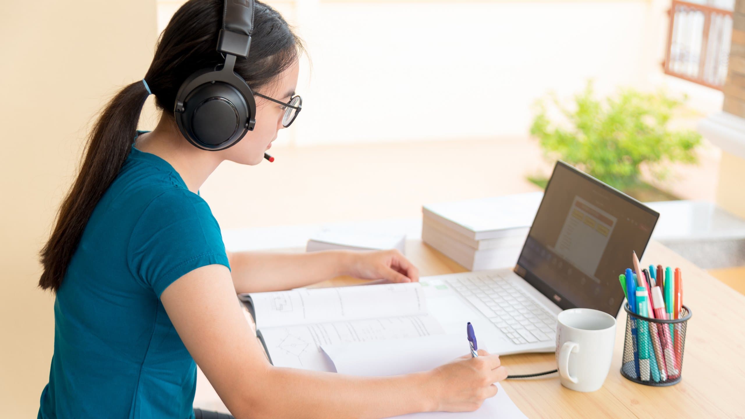 Learner with headphones taking notes beside a laptop